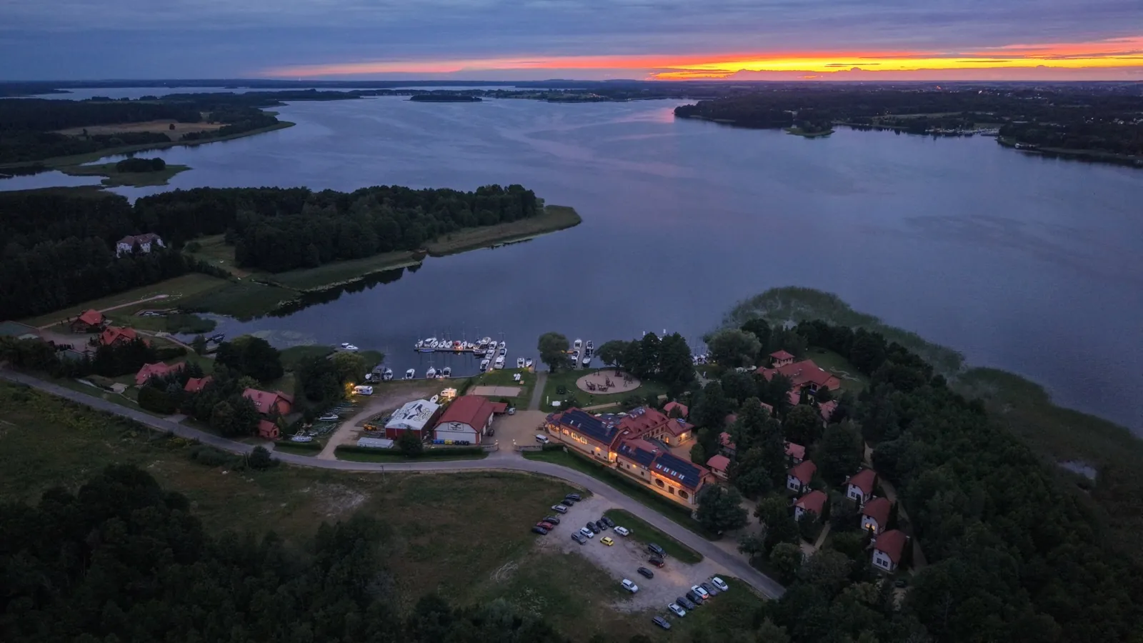 Mazurskie jezioro o zmierzchu z lotu ptaka: pomarańczowy pas nieba nad wodą i zabudowa nad brzegiem